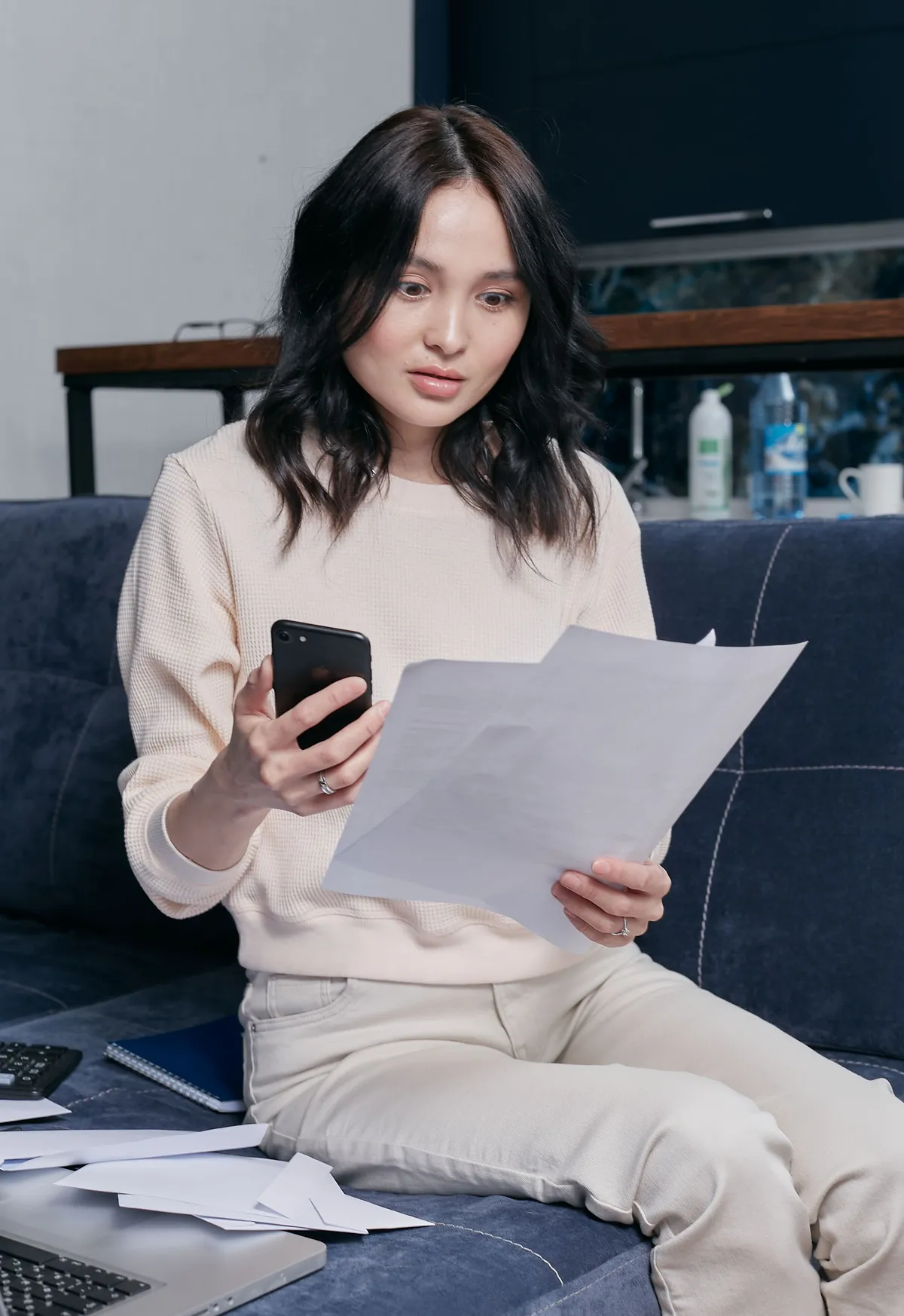 Mulher no sofá lendo um documento com o celular na mão (cena ilustrativa)